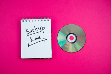Notepad with backup time text and dvd disk lie on a pink background.