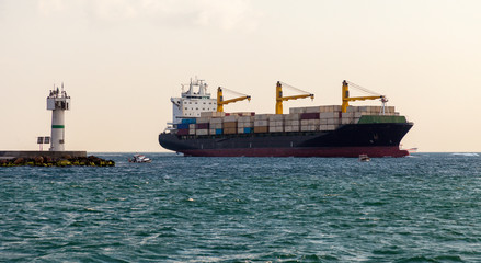 Huge cargo ship is passing by the lighthouse in Istanbul bosphorus. Cargo ship has 3 cranes on it and fully loaded. Lighthouse and cargo ferry next to each other with some little fishing boats nearby