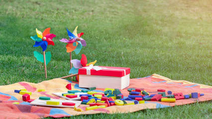 Fototapeta premium Colorful wooden blocks on the rug next to opened gift box and rainbow pinwheels on the grass in the garden or park.