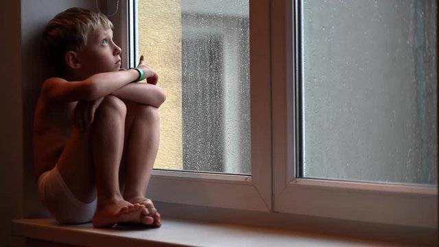 Little boy sitting on windowsill watching at raindrops on glass as it's raining outside. Loneliness, childhood, domestic life concept. Lockdown, isolation concept. COVID-19 concept.
