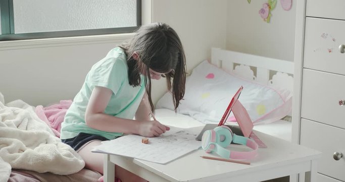 young girl home schooling or doing homework in her bedroom. She is writing in a notepad at a small table with a tablet computer and headphones on the table. She flicks her hair