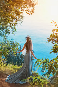 art fantasy photography. queen woman stands on river lake shore. sunset summer sun light. fashion model gray vintage long dress silhouette mermaid silver crown. concept divine freedom. Back rear view