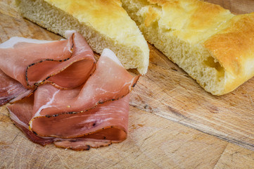 Sliced italian smoked speck ham with focaccia bread on a wooden cutting board close up.