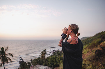 Girl in tattoos, stands on the edge of a cliff and looks through binoculars against the backdrop of the sea and sunset in Goa