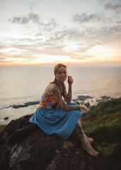 Fototapeta na wymiar Girl in tattoos, a multi-colored psychedelic T-shirt and a blue skirt sits on the edge of a cliff against the backdrop of the sea and sunset in Goa