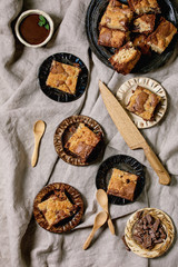 Trend baking Brookies chocolate brownies and cookies homemade cake sliced by squares in different ceramic plates, chocolate sauce, wooden spoons on grey linen cloth background. Flat lay, space
