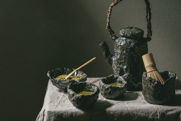 Japanese matcha green tea in craft ceramic cups with matcha powder, bamboo whisk, ceramic teapot on grey linen table cloth in dark room
