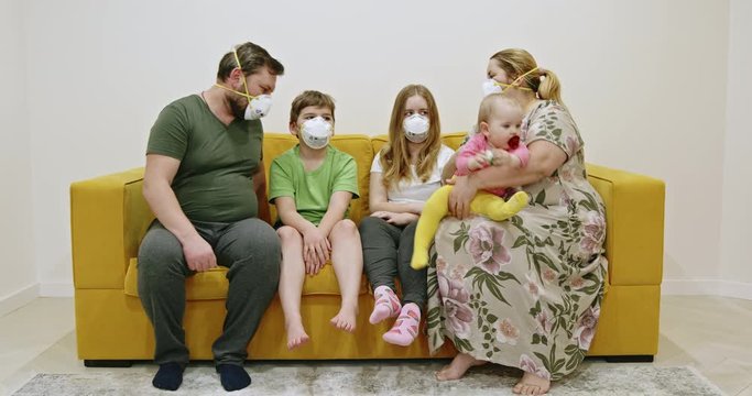 Family in masks sitting on sofa. Overweight mother and father in medical masks hugging kids and stacking hands together while sitting on yellow sofa during quarantine at home.