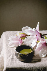 Japanese matcha green tea in wabi sabi ceramic cup with pink magnolia flowers on grey linen table cloth.