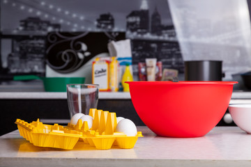 On the kitchen table, closeup is a red bowl, eggs in a yellow substrate, a glass cup on a background of ingredients for making dough or baking. A standard set of daily routine women.