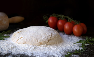 homemade pizza dough or pies on a dark rustic background, next to a branch of tomatoes and arugula greens and a rolling pin