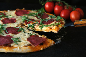 homemade pizza on a dark rustic background, next to a branch of tomatoes and arugula greens. A slice of pizza is cut off with a knife