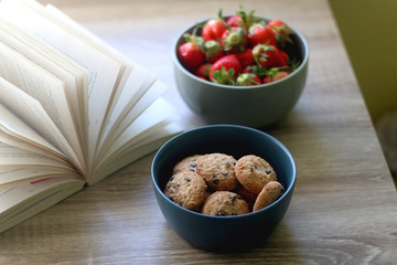 Bowl of chocolate chip cookies, bowl of strawberries and an open book on a table. Selective focus.