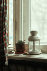 On a white wooden windowsill there is a candlestick-lantern with a burning candle and a green, home plant in a pot