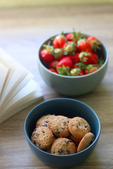 Bowl of chocolate chip cookies, bowl of strawberries and an open book on a table. Selective focus.