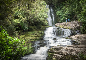 Landscape shot of big cascade waterfall in middle of forest with people included for scale. Shot made at McLean Falls in Catlins Forest Park, New Zealand