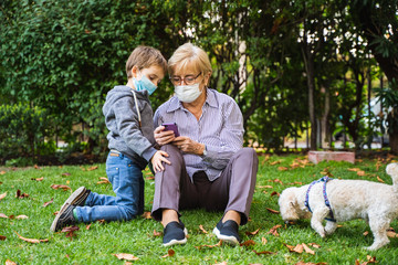 Obraz premium Grandmother and little kid playing with a smartphone in the backyard while wearing protective masks due to coronavirus outbreak
