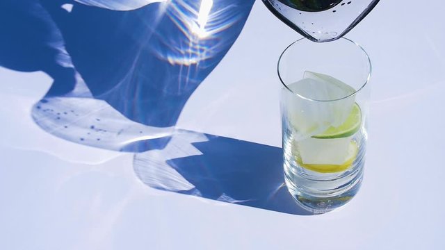Water poured from a glass jug into a glass with ice and lemon/lime.
Slow-motion. Hard light.