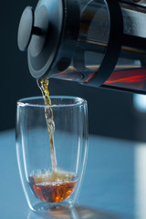 french press black tea pouring into a glass with a double bottom on a white table and grey background - vertical photo