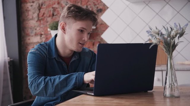 Young disabled man sitting in comfort wheelchair in modern apartment with bright and light room inside. Male using computer for chatting browsing internet. he smiles and laughs. having fun