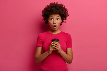 Waist up shot of emotional stupefied woman stares with bugged eyes, drinks coffee to go, realizes shocking news while talking with friend, enjoy hot drink, poses against pink studio background