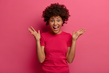 Emotive dark skinned woman has shocked cheerful expression, raises hands, shouts loudly, supports...