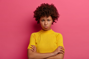 Discontent woman with sulking expression, keeps arms folded, looks angrily at camera, offended by hearing bad words, frowns face in displeasure, wears bright yellow clothes, isolated on pink wall