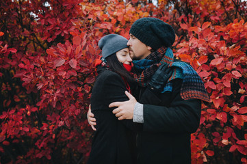 A young family couple in love in a coat, in hats and with scarves standing near the red autumn bushes and kiss. The concept of rest and relaxation in nature.