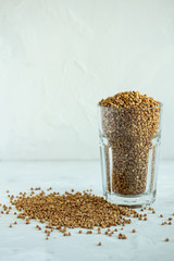 Close-up of buckwheat is scattered on a light gray table next to a transparent glass filled with buckwheat