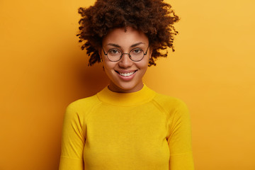 Portrait of lovely woman smiles gently, has curly hair, wears round transparent glasses and yellow jumper, looks directly at camera, listens pleasant news, poses indoor. Human face expressions