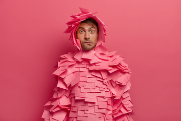 Puzzled young man bites lips and looks surprisingly at camera, reacts on astonishing news, makes funny costume of stickers, poses indoor against bright pink wall. Human facial expressions concept