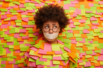 Photo of puzzled stressed Afro woman with mouth covered by tape, has no freedom of speech, being kidnapped, looks embarrassed aside, wears spectacles, poses over paper wall with adhesive notes
