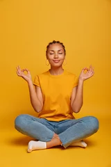Foto op Aluminium Peaceful calm dark skinned female model sits crossed legs on floor, practices yoga and tries to relax, breathes deeply, closes eyes, reaches nirvana, holds hands sideways, releases stress after work © Wayhome Studio