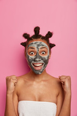 Joyous dark skinned woman applies beauty mask on face stares at camera, has broad smile clenches fists believes dreams come true, has bun hairstyle, isolated on pink wall. Wellbeing, skin care concept