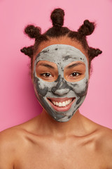 Close up shot of positive woman enjoys face treatment, applies clay mask, smiles broadly, has white teeth, stands naked alone, feels relaxed, isolated over pink background. Refreshing, spa, body care