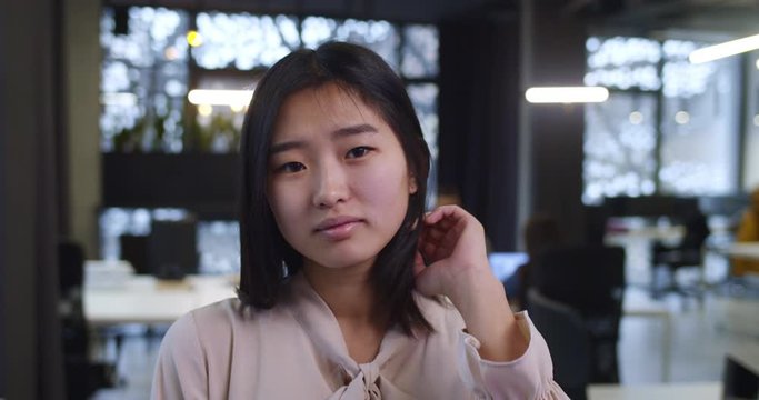 Close up of attracive happy young Asian woman looking at camera with smille and fixing her dark hair. Pretty girl standing in company office during workday. Female employee in firm.
