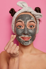Close up shot of beautiful Asian woman applies purifying black mask on face, has beauty treatments, makes korean like sign, wears grey headband, stands shirtless against pink background in bathroom