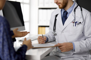 Unknown red-bearded doctor and patient woman discussing current health examination while sitting in sunny clinic, close-up. Medicine concept