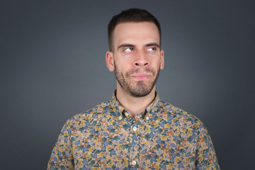 Gloomy, wondering young man face looking up lips together, being upset thinking about something important, keeps hands down, man standing indoors.