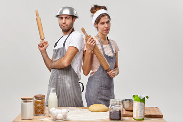 Serious woman and man cooks stand back to each other, hold rolling pins, take part in culinary battle, demonstrate cooking skills pose near table with ingredients at kitchen. We rule in culinary world