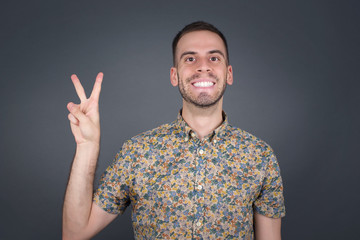 Young caucasian man standing against gray wall showing and pointing up with fingers number two while smiling confident and happy.