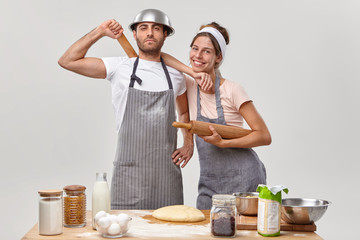 Time to cook. Friendly cook team make dough, hold wooden rolling pins, feel tired but satisfied, pose at kitchen near table with necessary ingredients. Woman and man participte in cooking contest