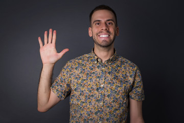 Young man standing against gray wall showing and pointing up with fingers number five while smiling confident and happy.