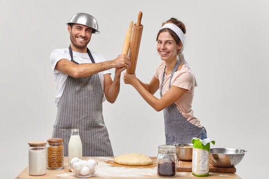 Professional restaurant competitors have battle with rolling pins, participate in culinary battle, look gladfully at camera, prepare tasty food, get ready for party weekend. Couple cooking together