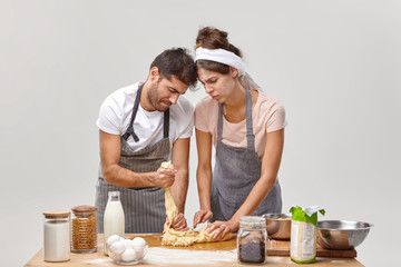 Horizontal shot of inexperienced couple prepare sticky dough for first time, being bad cooks, look awkwardly, wear aprons, stand near table with products. Kitchen disaster and cooking failure