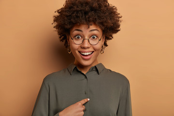 Am I what you need? Happy surprised female model points at herself, asks who me, smiles broadly, looks through round glasses, being chosen in business team, has positive reaction on hearing her name