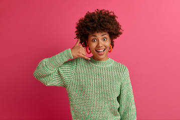 Positive talkative mixed race woman makes call me gesture, keeps contact with friends on self isolation over telephone, smiles pleasantly, poses well dressed indoor, tries to gets someones number