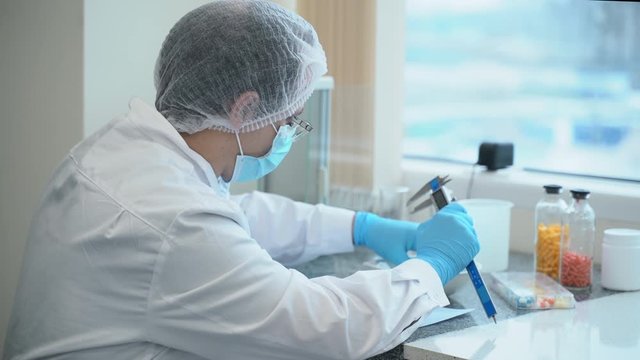 Processional man is examining new drug and writing document at table in pharmaceutical lab spbas. Male scientist measures white pills using tool and takes notes while sitting at desk in modern company