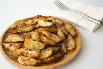 Oven-baked potato wedges with dried herbs on a wooden plate with a napkin and a fork in the background