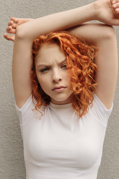 The portrait of the curly red hair girl with heart shaped fake freckles staying near the wall on the street with her hands up and angree face in a white t-shirt